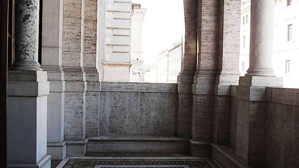 Loggia in pietra di un edificio storico con colonne e capitelli decorati, soffitto a cassettoni e parapetto alto. Il pavimento è rivestito da un mosaico geometrico a rombi con bordura ornamentale, affacciato sulla strada e su altri edifici.