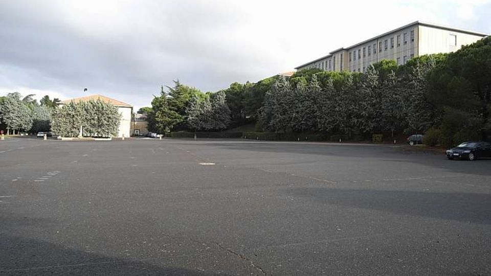 Ampio piazzale asfaltato quasi vuoto, affiancato da una fascia di alberi e vegetazione su un pendio. Sullo sfondo si vedono un edificio basso a sinistra e un lungo edificio rettangolare a più piani sulla destra, sotto un cielo nuvoloso.