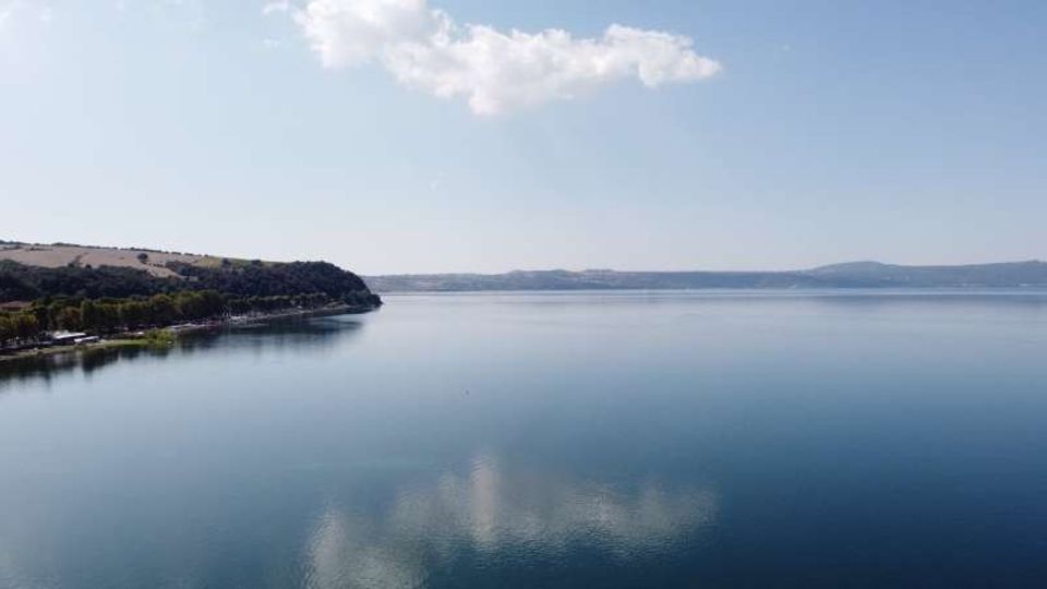 Ampio lago dalle acque calme con riflessi di nuvole, visto da lontano. Sulla sinistra si vede una costa boscosa con una linea di alberi, mentre all’orizzonte compaiono colline e una sponda distante sotto un cielo sereno.
