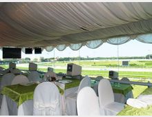 Sala hospitality con soffitto drappeggiato in tessuto e tavoli apparecchiati, con sedie rivestite di bianco e tovaglie verdi. Grandi vetrate affacciano su un ippodromo con pista e prato, con schermi appesi e piccoli monitor lungo il parapetto.
