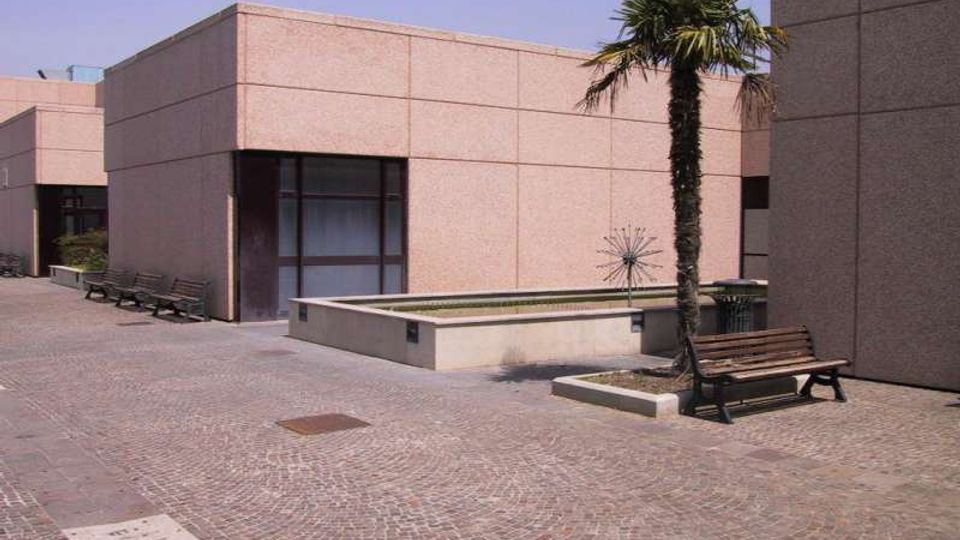 Cortile esterno pavimentato in pietra tra edifici moderni rivestiti in pannelli chiari, con grandi finestre scure. Al centro c’è una vasca rettangolare rialzata e una palma, con panchine lungo il perimetro.