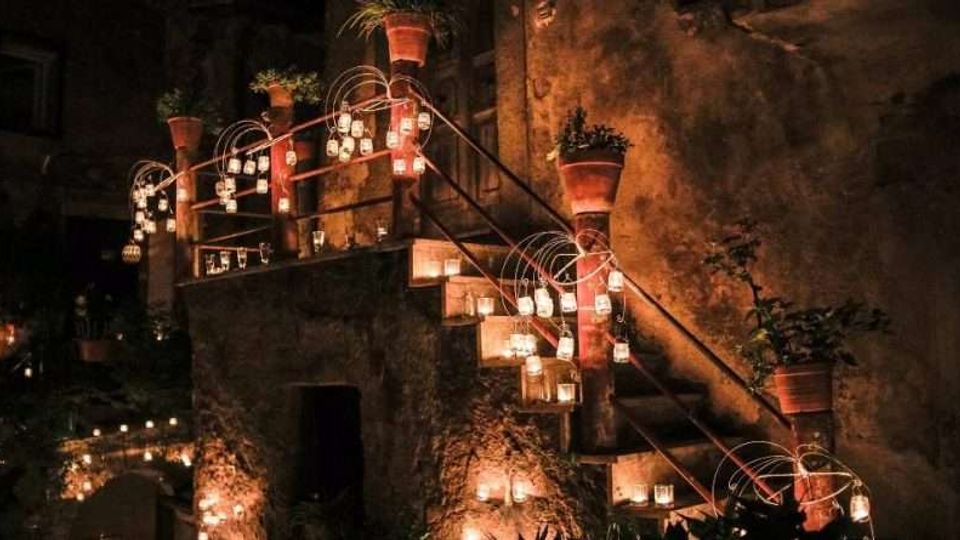 Scalinata esterna in pietra con corrimano in ferro, decorata con file di piccole lanterne luminose appese e poggiate sui gradini. Lungo il percorso ci sono colonne con vasi di terracotta e piante, addossate a una parete in pietra illuminata in modo soffuso.