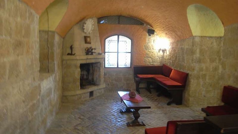 Salotto rustico in pietra con soffitto a volta in mattoni, pavimento in mattoni a spina di pesce e camino in muratura. Al centro c’è un tavolino basso, con panche e divani imbottiti rossi lungo le pareti e una finestra ad arco sul fondo.