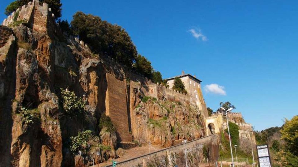 Strada ai piedi di una parete rocciosa scoscesa con vegetazione, sormontata da resti di mura in pietra e da un edificio storico. In basso si vede un parcheggio con segnaletica e un piccolo arco d’ingresso lungo il percorso.