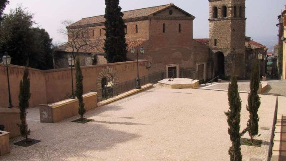 Piazza lastricata in pietra con aiuole e cipressi, circondata da muretti e lampioni. Sullo sfondo si vede una chiesa in mattoni con campanile in pietra e tetti in coppi, affacciata su un leggero pendio urbano.