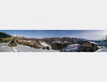 Panorama montano innevato con vallate e crinali sullo sfondo, sotto un cielo limpido. In primo piano si vedono case e recinzioni su un pendio, con neve sui tetti e piccoli alberi sparsi.