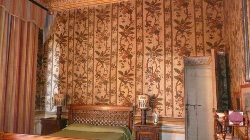 Camera da letto in stile classico con letto in legno e comodini, pareti rivestite da carta da parati a motivi floreali verticali e soffitto decorato con cornici ornate. Sulla sinistra si vede una tenda a righe davanti a una finestra e sulla destra una porta interna.