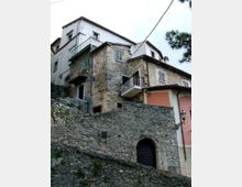 Edifici residenziali su pendio con facciate in pietra e intonaco, tetti in tegole e piccoli balconi con ringhiere metalliche. In primo piano si vede un alto muro di contenimento in pietra con arco e portone, affiancato da una lanterna a muro.