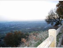 Belvedere su una collina con staccionata in legno in primo piano, affacciato su una vasta valle con campi e piccoli insediamenti. Vegetazione e alberi ai lati incorniciano l’orizzonte lontano sotto un cielo chiaro.