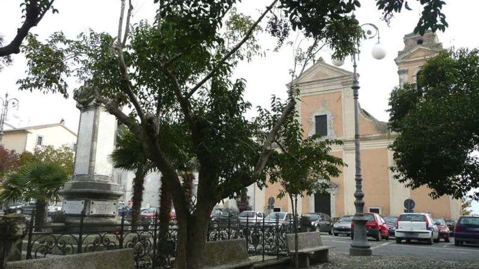 Piazza lastricata in pietra con alberi e panchine in primo piano, delimitata da una ringhiera in ferro e con un lampione. Sullo sfondo si vede una chiesa con facciata color pastello e campanile, affiancata da auto parcheggiate e da un monumento in pietra.