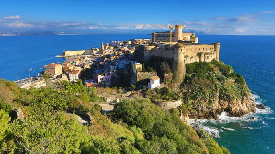 Veduta panoramica di un borgo costiero su un promontorio roccioso, dominato da una grande fortezza in pietra con bastioni e mura. Intorno si vedono case compatte e vegetazione mediterranea, con il mare blu che circonda la scogliera e l’orizzonte sullo sfondo.