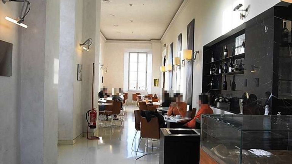 Interno di un ristorante o caffetteria in stile moderno, con lungo corridoio, pavimento chiaro lucido e soffitto alto. A destra si vede un bancone con vetrina e scaffali con bottiglie, mentre in fondo ci sono tavoli con sedie e una grande finestra che illumina la sala.