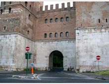 Ingresso ad arco in una cinta muraria di un edificio storico in mattoni con merli e piccole finestre, poggiato su una base in pietra chiara. Davanti passa una strada asfaltata con segnaletica di divieto e un semaforo.