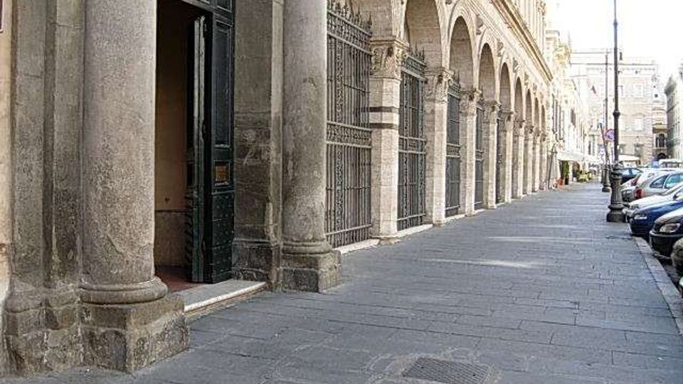 Portico urbano lungo una strada, con grandi colonne in pietra e una sequenza di archi che sostengono la facciata di un edificio storico. Sul marciapiede ampio si vedono cancellate metalliche, un ingresso con porta aperta e auto parcheggiate sul lato della carreggiata.