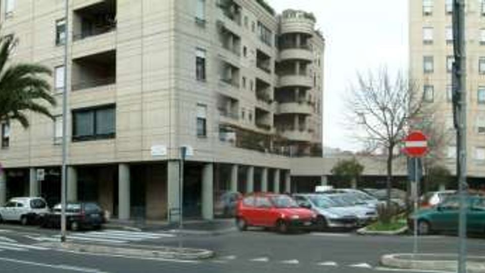 Ampia strada urbana con incrocio e strisce pedonali, fiancheggiata da palme e palazzi residenziali multipiano con balconi. Lungo i marciapiedi sono presenti auto parcheggiate e segnaletica stradale, tra cui cartelli di divieto di accesso.