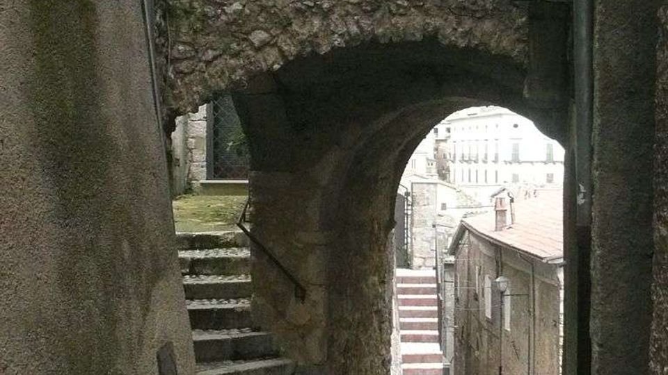Vicolo stretto in un centro storico, con pavimentazione in ciottoli e un arco in pietra grezza che collega due edifici. A sinistra ci sono gradini in pietra con corrimano, mentre oltre l’arco si intravede una strada in discesa con case e scalinate.