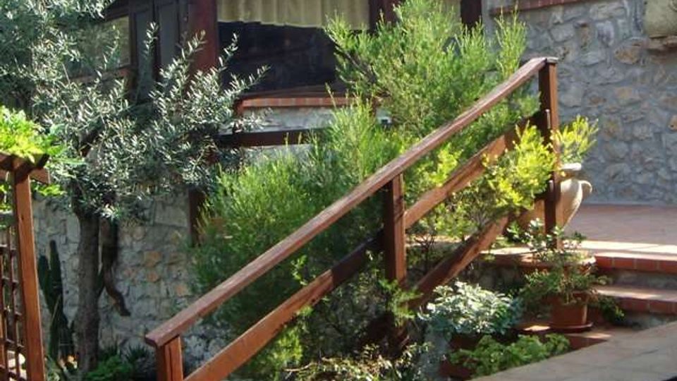 Casa con veranda in legno e tetto spiovente, costruita su un terrazzamento con muri in pietra. In primo piano una scalinata con ringhiera in legno e vasi di piante, circondata da vegetazione e alberi.