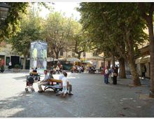 Piazza urbana lastricata con ampi alberi che creano ombra, panchine e una struttura a forma di chiosco vetrato al centro. Sullo sfondo si vedono edifici bassi con portici e ombrelloni di bar e ristoranti all’aperto.