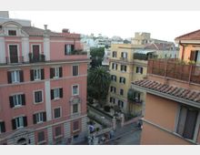 Vista dall’alto di un quartiere urbano con palazzi residenziali color rosa e giallo, tetti in coppi e persiane scure. Tra gli edifici si vede una strada e un piccolo giardino con una grande palma e vegetazione.