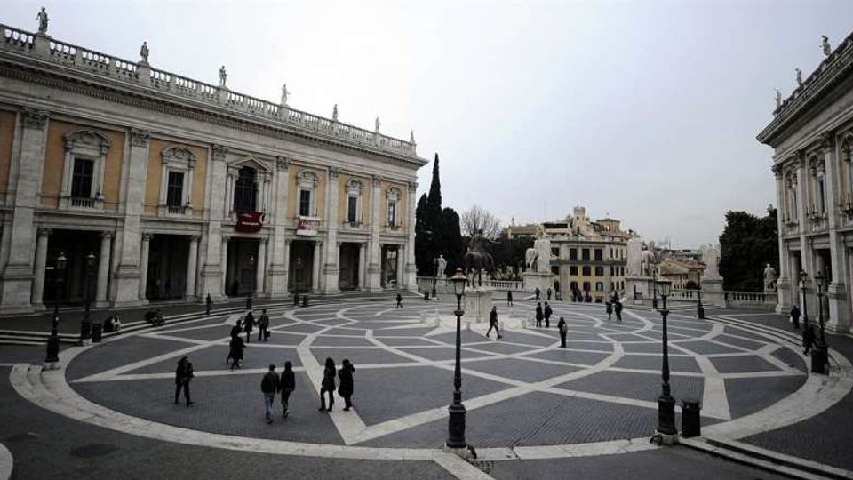 Ampia piazza pavimentata con disegni geometrici in pietra, circondata da edifici storici neoclassici con portici, colonne e balaustre con statue. Al centro e ai lati si vedono lampioni in ferro e alcune alberature, con altri palazzi sullo sfondo.