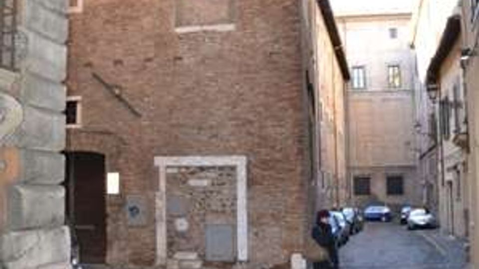 Vicolo urbano lastricato con un grande edificio in mattoni a vista sulla sinistra, con finestre piccole e una porta murata. La strada stretta prosegue tra palazzi storici con un lampione sospeso e alcune auto parcheggiate sullo sfondo.