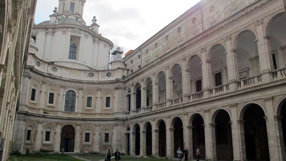 Cortile interno di un complesso storico in pietra, con lunghe gallerie porticate ad archi su due livelli e una facciata curva con grandi finestre. Sullo sfondo si vede una cupola con lanterna e decorazioni barocche, con prato e pavimentazione in pietra al centro.