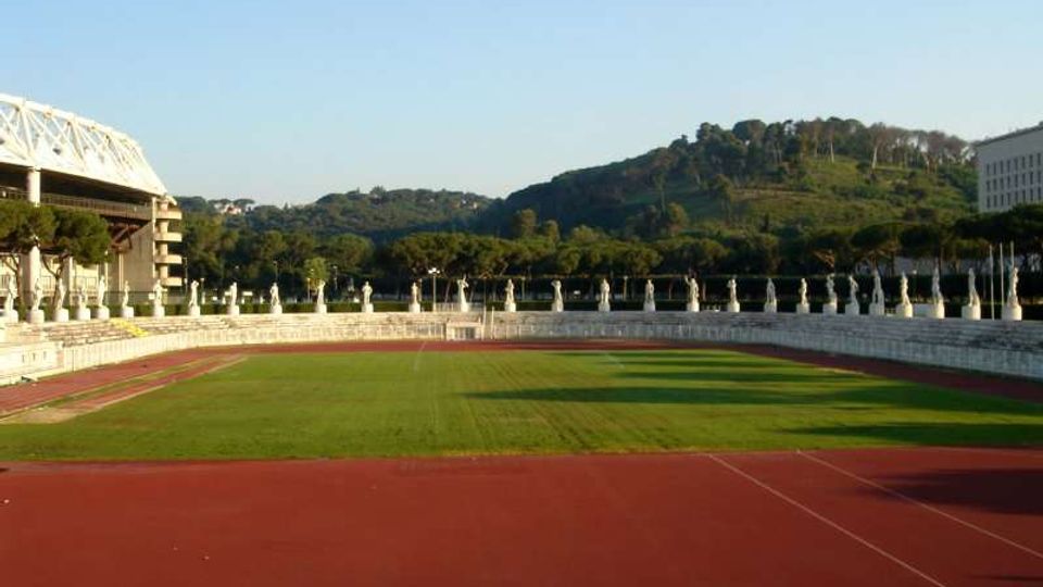 Vista di uno stadio con pista di atletica rossa e campo erboso centrale, circondato da gradinate basse e una fila di statue lungo il bordo. Sullo sfondo si vedono colline alberate e alcuni edifici ai lati.