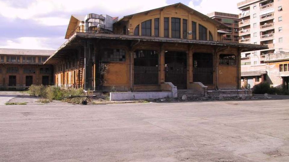 Grande edificio industriale in mattoni, con ampie finestre ad arco e una tettoia sporgente sostenuta da colonne, affacciato su un vasto piazzale asfaltato. Sullo sfondo si vedono altre strutture del complesso e palazzi residenziali moderni.