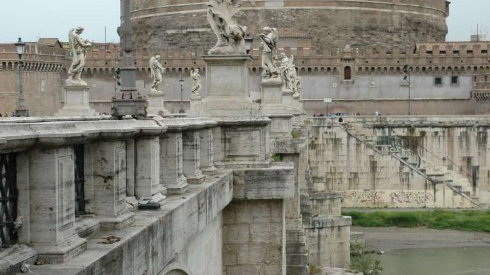 Ponte in pietra con parapetto decorato da statue e lampioni che attraversa un fiume, visto in prospettiva lungo le arcate. Sullo sfondo si innalza una grande fortezza circolare in muratura con edificio superiore e statua sulla sommità.