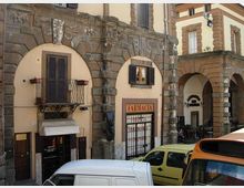 Facciata di un edificio storico con grandi archi in pietra e pareti intonacate, con finestre a persiane e un piccolo balcone in ferro battuto. Al piano strada si vede l’ingresso di una farmacia con insegna “FARMACIA” sotto un portico ad archi; in primo piano sono parcheggiati alcuni veicoli.