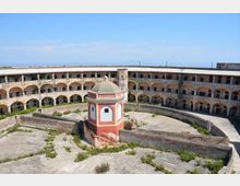Cortile interno circolare di una grande struttura in muratura con due ordini di arcate e corridoi perimetrali. Al centro si trova un piccolo edificio ottagonale rosso e bianco con cupola, circondato da pavimentazione in cemento con vegetazione spontanea.