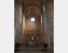 Interno di una chiesa in pietra con alta navata e volte a crociera, colonne massicce e abside con altare rialzato. Sul fondo si vedono un rosone circolare in alto e tre strette finestre ad arco con vetri colorati.