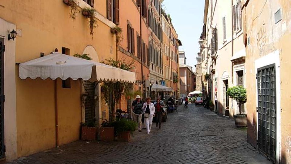 Strada stretta in ciottoli tra edifici alti dai colori ocra e arancio con persiane scure, con tende bianche e fioriere lungo il lato sinistro. La via prosegue in prospettiva con lampioni a parete, portoni e finestre sui fronti degli edifici.