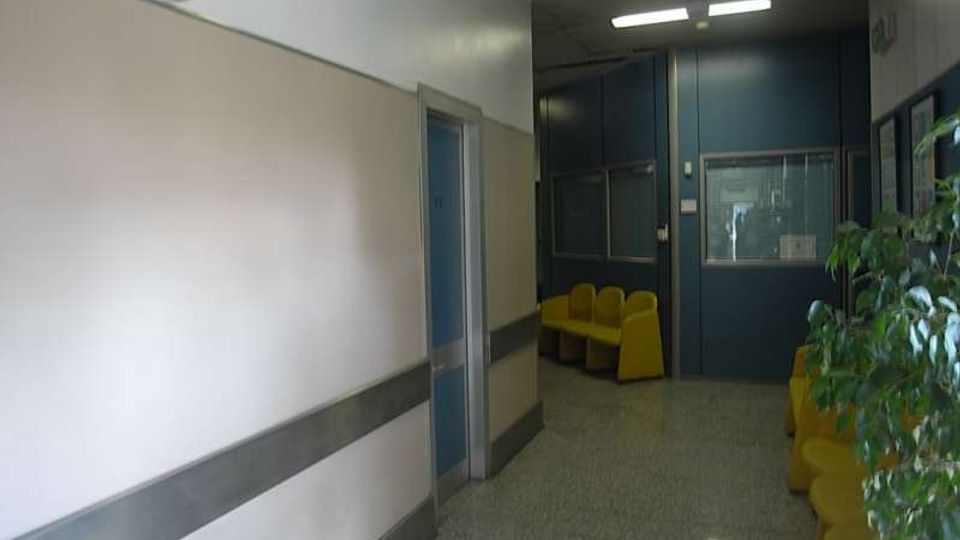 Corridoio interno di una struttura pubblica con pavimento in graniglia, pareti chiare con fascia grigia e orologio a parete. In fondo si vede una zona d’attesa con sedute gialle, pannelli e porte blu, illuminazione a neon e una pianta lungo il lato destro.