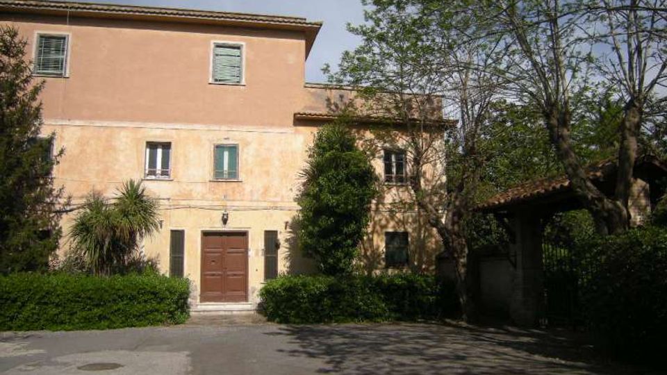 Edificio a due piani intonacato color pesca con tetto in tegole e porta d’ingresso centrale, affacciato su un cortile asfaltato. Davanti ci sono siepi e alberi, con un passaggio coperto sul lato destro accanto a una recinzione.