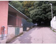 Strada asfaltata in salita che costeggia un edificio in mattoni con recinzione metallica e piccolo accesso, affiancata da un muro e un lampione. In fondo si intravede un’auto parcheggiata davanti a un’area alberata fitta.