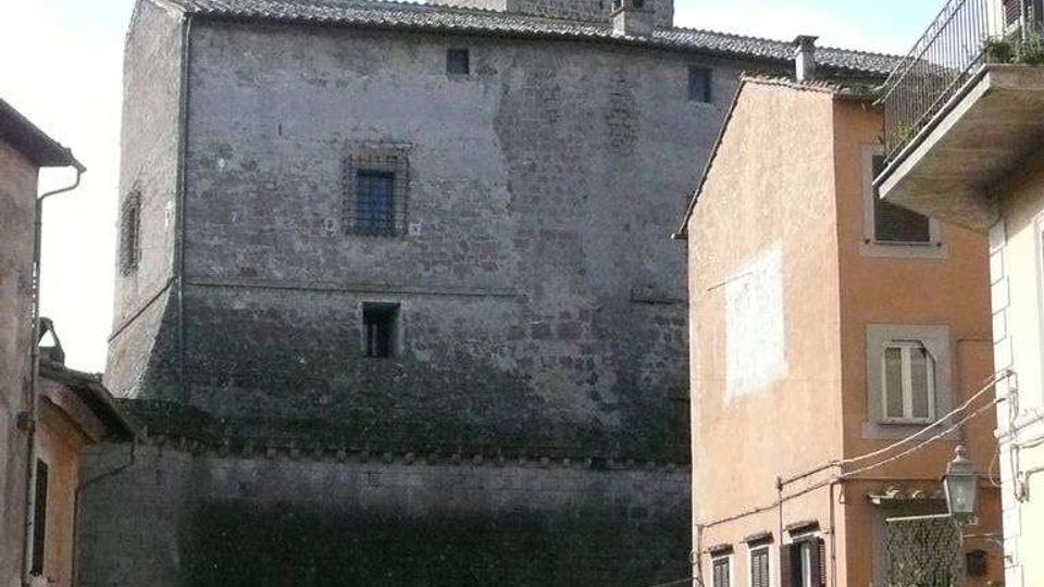 Edificio storico in pietra simile a una piccola fortezza, con alte mura quasi prive di aperture e una torre quadrata che svetta sopra il tetto. In primo piano si vede una strada stretta tra case residenziali con balconi e alcune auto parcheggiate.