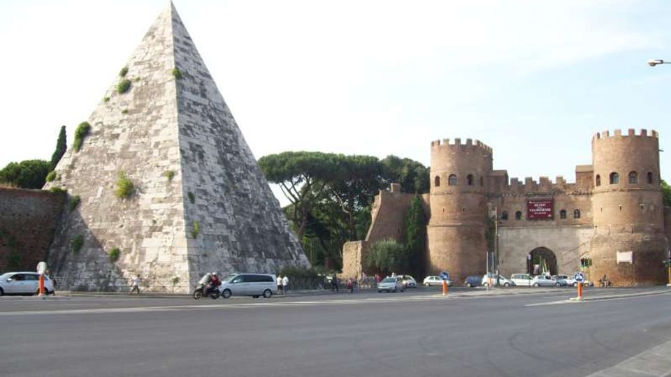 Grande piramide in pietra accanto a una strada ampia, con vegetazione sparsa sulle superfici. Sullo sfondo si vede una porta fortificata in mattoni con due torri cilindriche merlate e alberi a chioma larga dietro le mura.