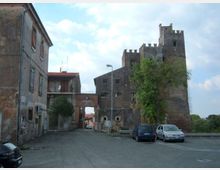 Strada di un borgo con edifici in muratura e un arco di passaggio, affiancata da un grande edificio fortificato in mattoni con torri merlate. Sul lato destro si vedono alberi e alcune auto parcheggiate lungo il bordo della carreggiata.