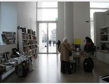 Interno di un negozio di libri o spazio espositivo moderno con soffitti alti, scaffali e tavoli pieni di volumi e riviste. In fondo, una grande porta vetrata a tutta altezza si apre su una piazza esterna con edifici visibili.