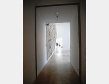Corridoio interno di un edificio moderno con pareti bianche e pavimento in pietra chiara, illuminato da faretti incassati nel soffitto. Sul lato sinistro si vede una grande parete decorata con disegni lineari, mentre in fondo si apre un ambiente più ampio e luminoso.