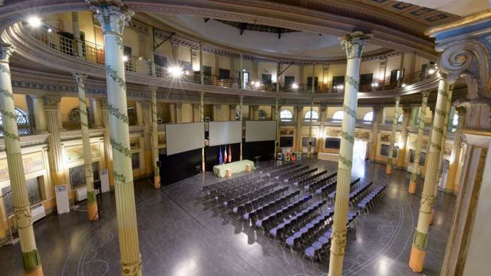 Ampia sala conferenze in un edificio storico con pianta ovale, colonne decorate e balconata perimetrale a due livelli. Al centro ci sono file di sedie rivolte verso un palco con schermi e tavolo, illuminate da luci a soffitto.