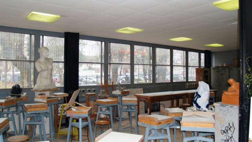 Aula-laboratorio con numerosi sgabelli e piccoli banchi da lavoro, tavoli lunghi e un busto in gesso su piedistallo. Lungo una parete ci sono grandi finestre a nastro con inferriate che affacciano sull’esterno, e un controsoffitto con plafoniere rettangolari.