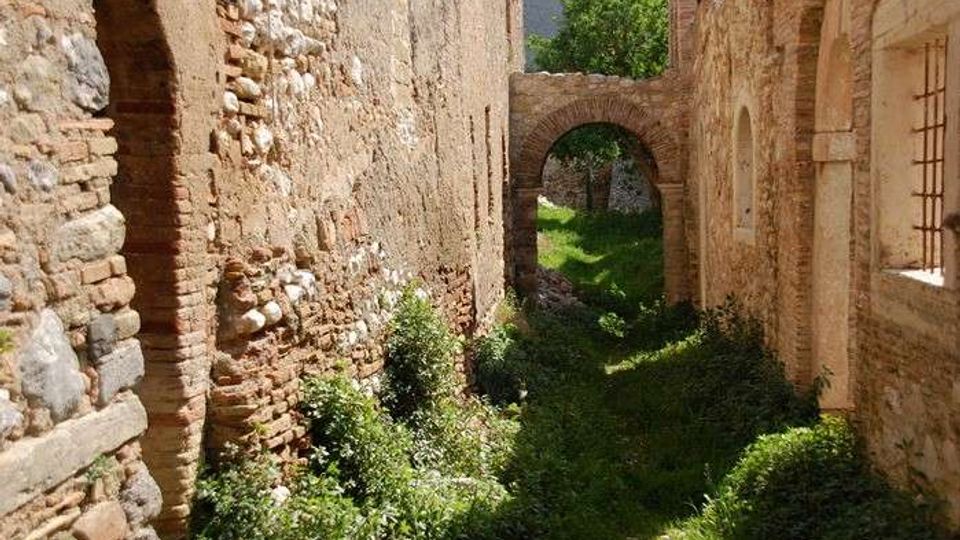 Passaggio stretto tra alti muri in pietra e mattoni di un edificio antico, con vegetazione che cresce sul terreno. In fondo si vede un arco in muratura che conduce a un’area verde illuminata dal sole.