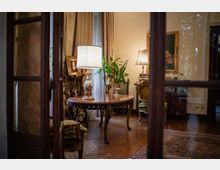 Interno di un salotto classico visto attraverso una porta a vetri, con pavimento in parquet e tappeto. Al centro c’è un tavolo rotondo intagliato con lampada, circondato da poltrone imbottite, quadri incorniciati, tende pesanti e piante vicino alla finestra.