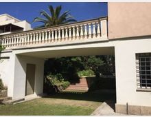 Cortile di una casa con prato e un portico passante sotto una terrazza con balaustra bianca, affiancato da pareti chiare e una finestra con grata. Sullo sfondo si vedono una scala in mattoni che sale tra vegetazione e una palma alta.