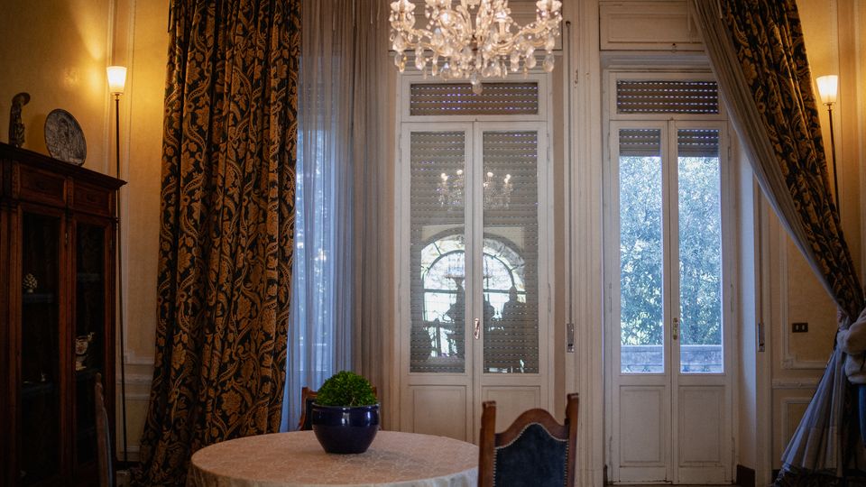 Interno di una sala da pranzo in stile classico con lampadario in cristallo, tavolo rotondo e mobili in legno. Grandi porte-finestre bianche con tende pesanti e leggere si affacciano su un balcone con alberi visibili all’esterno.