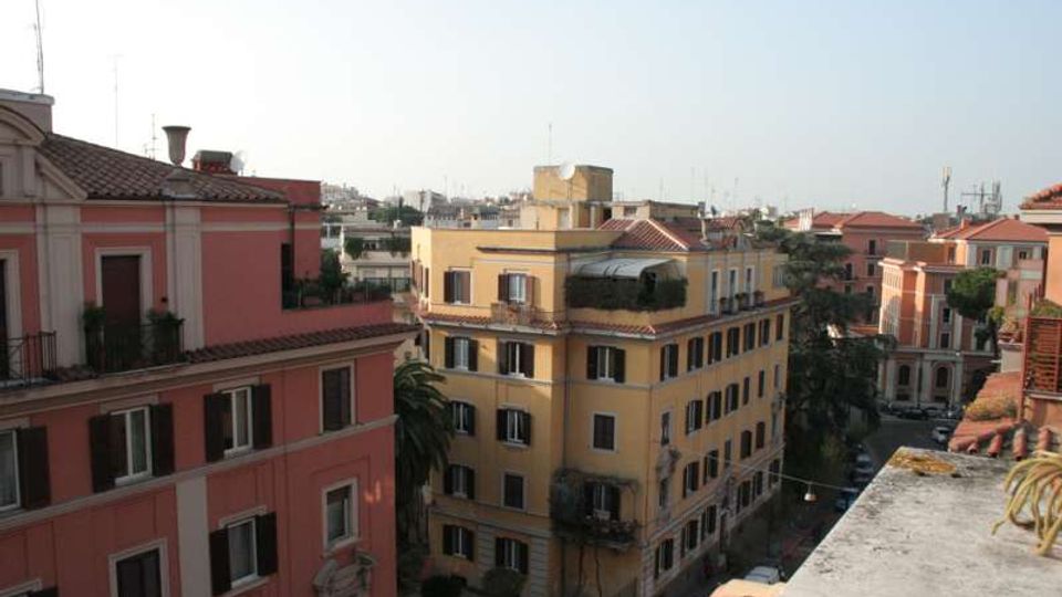 Veduta urbana dall’alto con palazzi residenziali in stile tradizionale dai toni rosa e giallo, con tetti in tegole, balconi e finestre con persiane. Sullo sfondo si vede un quartiere fitto di edifici e una strada alberata tra i palazzi.