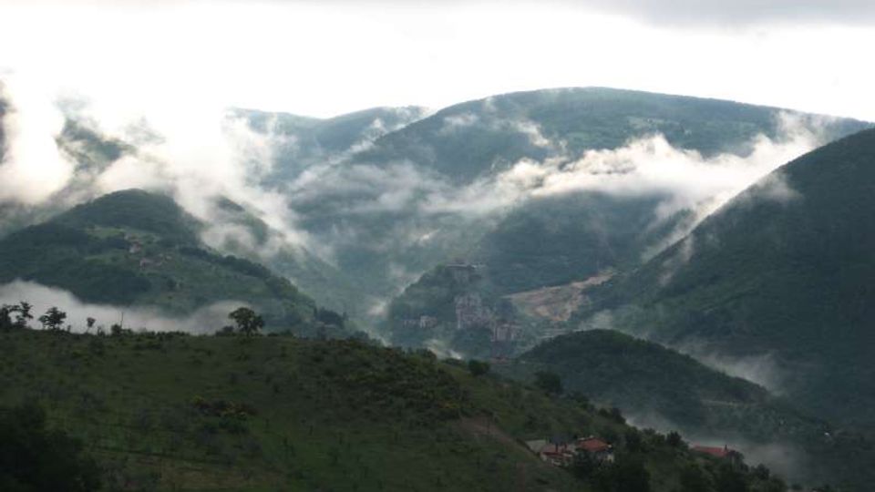 Panorama di una valle montana con colline e montagne verdi, attraversata da banchi di nebbia e nuvole basse. In primo piano si vedono pendii erbosi con pochi alberi e alcune case sparse.