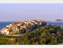 Veduta panoramica di un borgo medievale su un crinale, con case in pietra addossate tra loro e una torre campanaria, circondato da colline verdi. Sullo sfondo si vede il mare con una costa lontana sotto un cielo sereno.